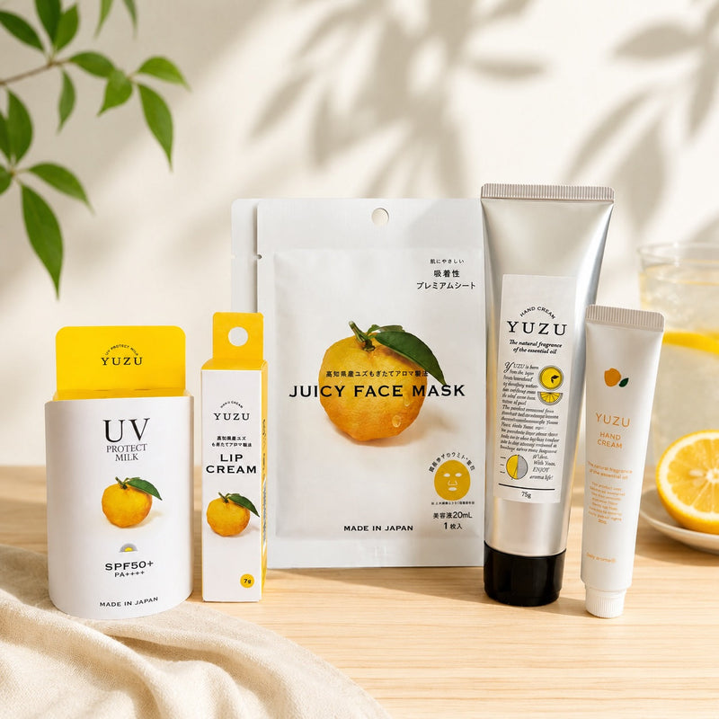 A YUZU skincare set arranged on a light wood surface with soft leaf shadows. The set includes a Juicy Face Mask sheet mask, a UV Protect Milk SPF50+ bottle, a Lip Cream tube, and a Lip Crème.