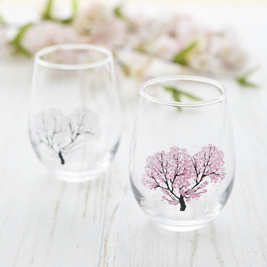 Two stemless glasses with a cherry blossom design that changes color when cold beverages are poured into them, presented on a white surface with cherry blossoms in the background.