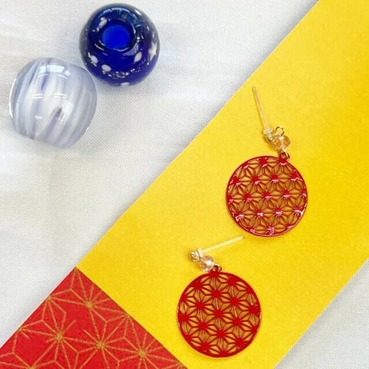 Red hemp leaf earrings on a yellow and red fabric background with blue and white marbled objects.