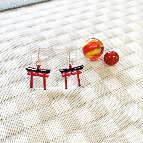 A pair of Torii gate-shaped earrings in vermilion and black, displayed alongside a red and yellow object on a textured background.