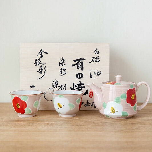 A set of Japanese ceramic teaware with hand-painted camellia flowers, including a teapot and two cups, displayed in front of a gift box with calligraphy.