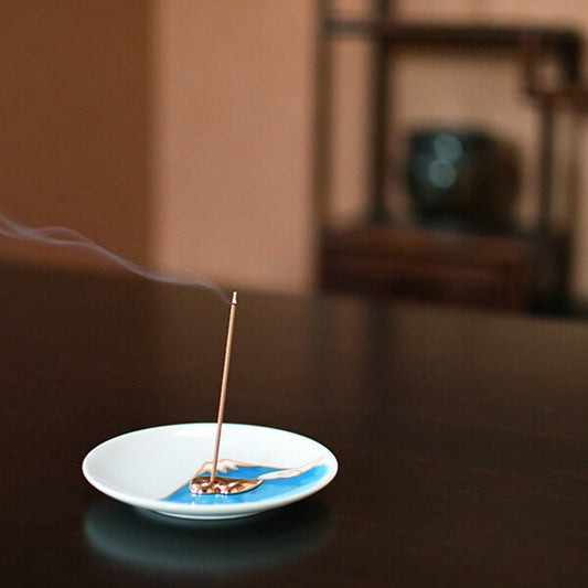 An incense stick is burned on a ceramic plate with a blue and white design, emitting smoke, placed on a table with a blurred background.