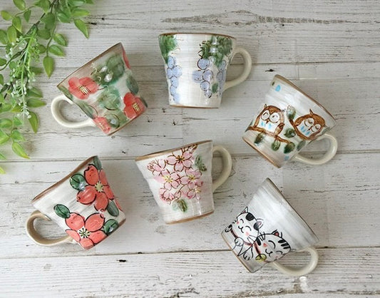 A collection of hand-painted Mino-yaki ceramic mugs with various patterns, including floral and animal motifs, displayed on a woven mat.