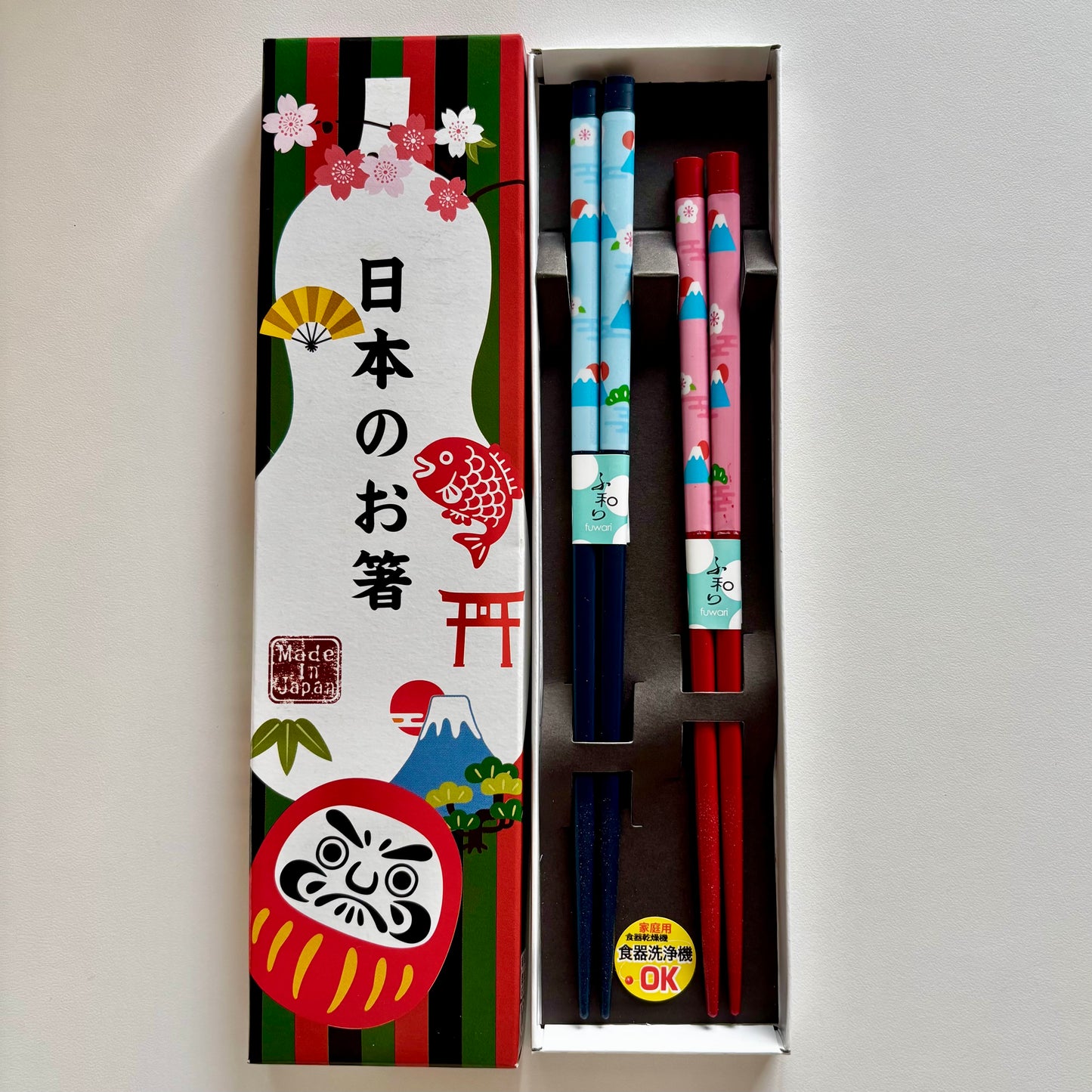 Pair of Japanese chopsticks in red and navy with Mt. Fuji, daruma, and cherry blossom motifs, presented in a colorful paper gift box