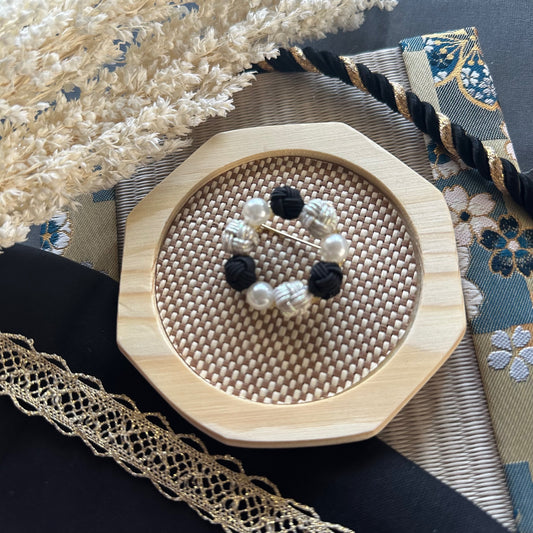 Handcrafted black Mizuhiki brooch with pearl-like beads displayed on a woven mat