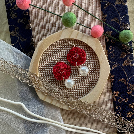 Red Mizuhiki knot earrings with pearl accents on a wooven mat with decorative background