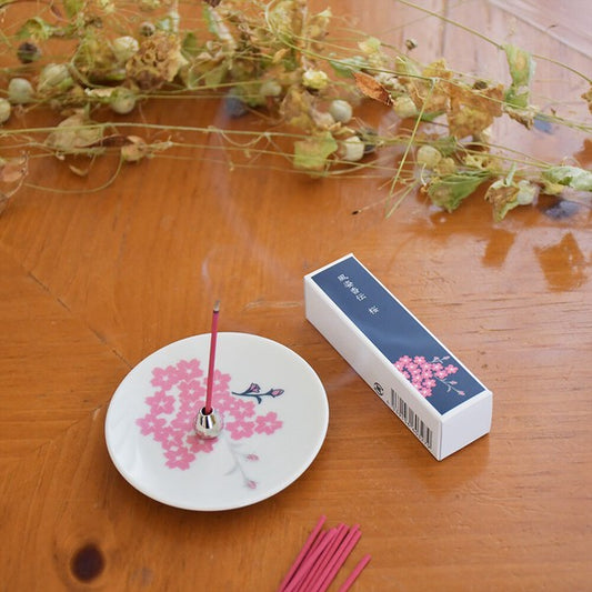 Sakura incense burning on silver-toned holder with ceramic plate, releasing floral fragrance