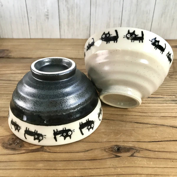 A pair of ceramic bowls with black cat designs on them, one bowl is black with a white interior and the other is beige with a gray interior, both displayed on a wooden surface.