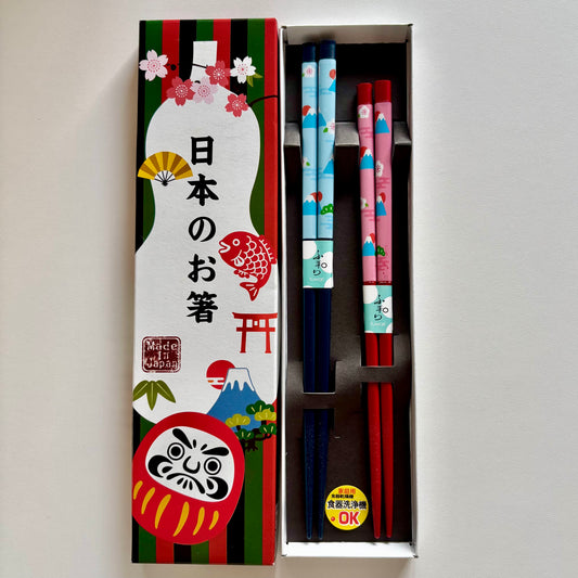 Pair of Japanese chopsticks in red and navy with Mt. Fuji, daruma, and cherry blossom motifs, presented in a colorful paper gift box