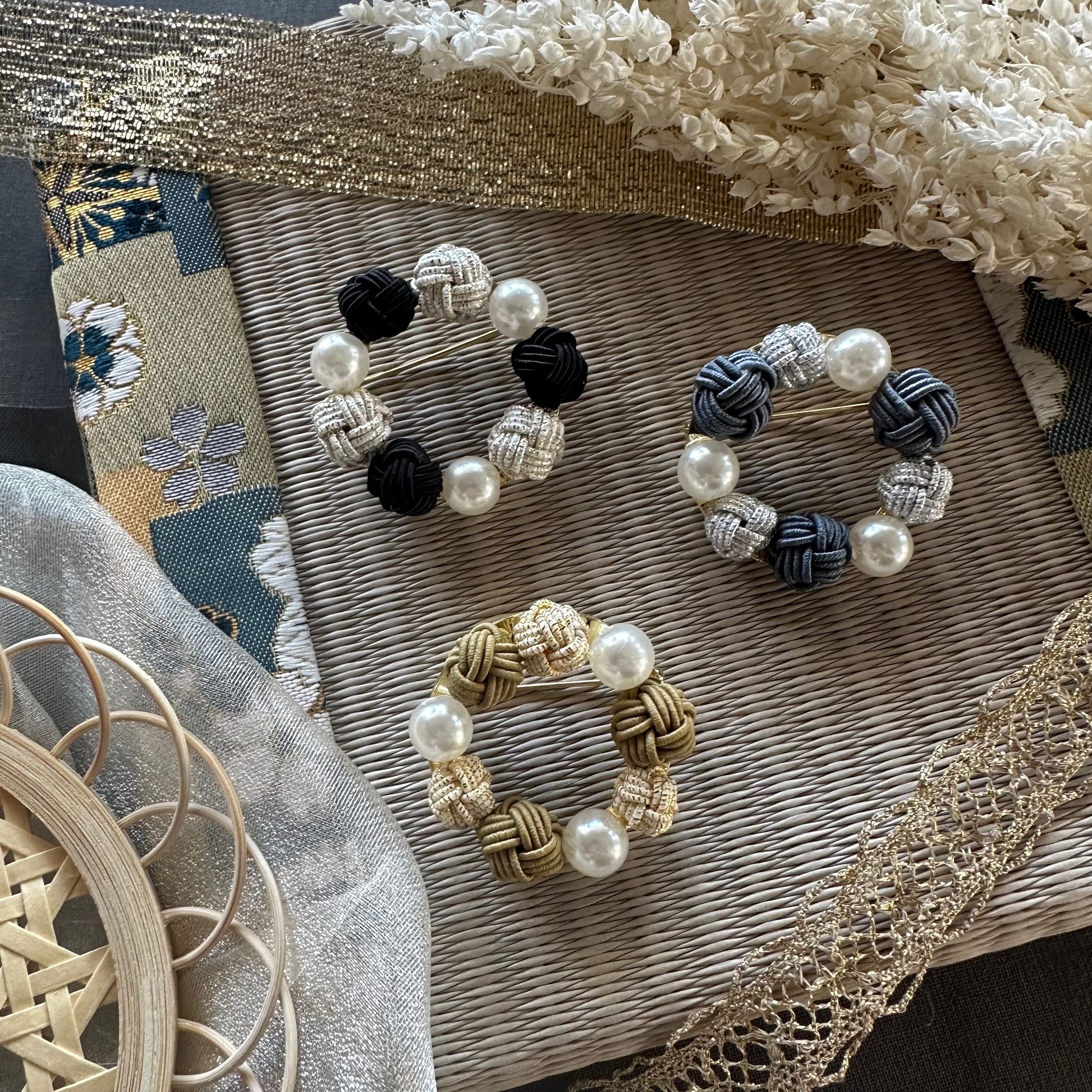 A collection of three handcrafted Mizuhiki Pearl Brooches in black, white, and gold colors, displayed on a woven mat.