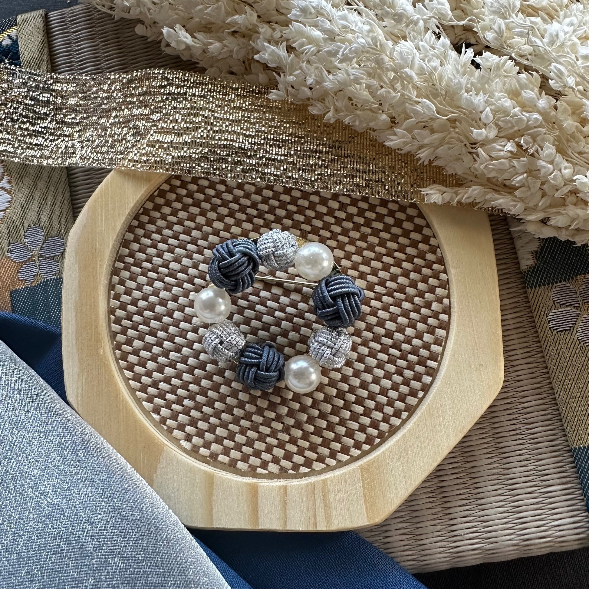 Handcrafted grey Mizuhiki brooch with pearl-like beads displayed on a woven mat

