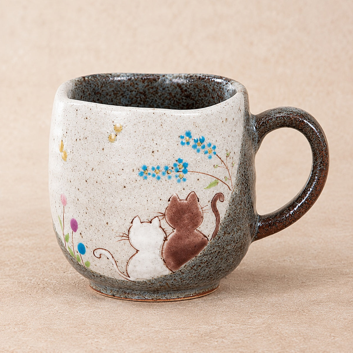 Handmade kutani-ware ceramic mug with a speckled beige and gray exterior, featuring a painted illustration of two small cats and blue flowers, set against a neutral beige background.